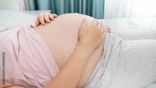 Closeup of young pregnant woman lying on bed at hospital room and stroking her big belly before giving birth. Concept of pregnancy, preparing and expecting child.