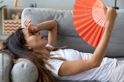 Close up overheated woman waving orange paper fan, lying on couch at home, exhausted sick young female feeling unwell, suffering from heating, fever or hot summer weather, touching forehead photo