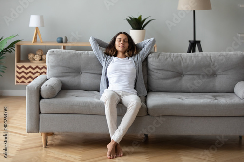 Full length peaceful woman with closed eyes resting on couch, leaning back with hands behind head, enjoying lazy weekend at home, calm young female relaxing, daydreaming, taking nap on sofa