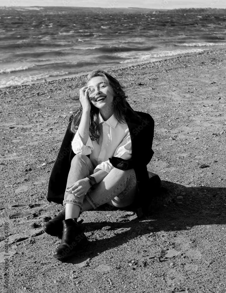 Portrait of a laughing young woman in black jacket, white shirt and jeans enjoying of sunlight sitting on beach sand in front of sea. Girl having fun outside. Urban style and fashion. Casual outfit