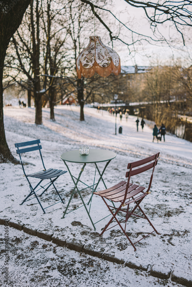 Outdoor cafe with sitting chairs and lamp over metal table with the people walking in the background at Sagene in Oslo, Norway during winter and snow