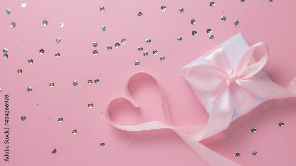 Gift box, heart shaped ribbon and shiny confetti on a pink background.