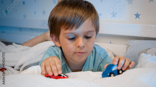 Smiling little boy playing with two toy cars while lying in bed at night. CHild playing before going to sleep.