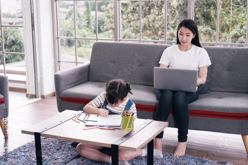 Smiling Asian mother and little asian girl child is drawing and Painting with wooden colored pencils on paper. Mother is online working with laptopin living room. Homeschool and educational concept photo