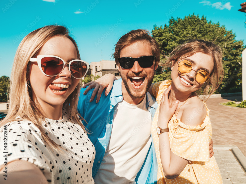 Group of young three stylish friends in the street.Man and two cute female dressed in casual summer clothes.Smiling models having fun in sunglasses.Women and guy making photo selfie on smartphone