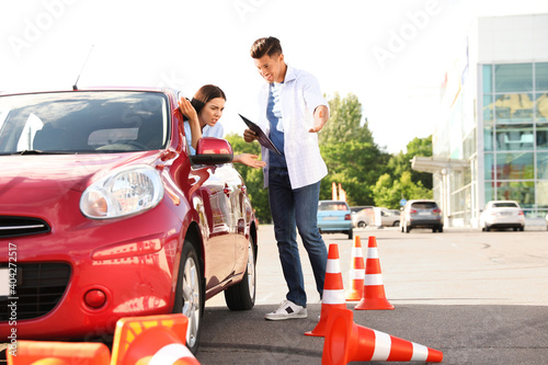 Stressed young woman in car near instructor and fallen traffic cones outdoors. Failed driving school exam © New Africa
