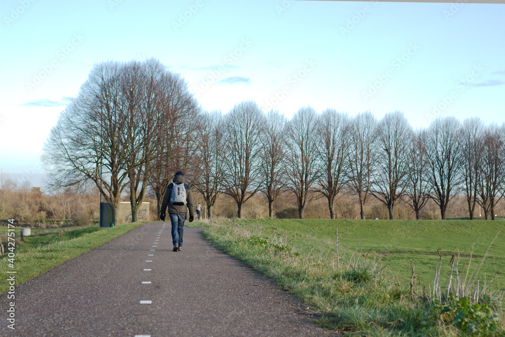 A man walking on a road with a backpack with a row of trees and green meadow. Outdoor hiking activities for healthy living concept.