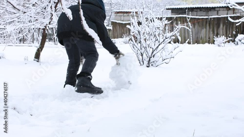 A little kid boy in dark clothes rolls a big snowball for showman on white snow at winter day, side view