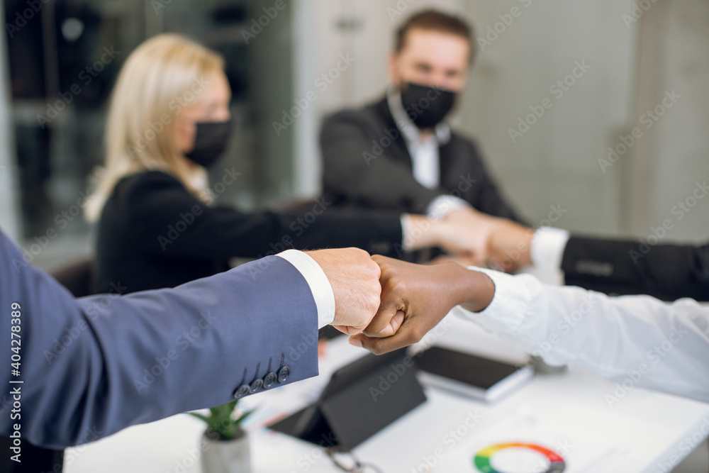 Close up cropped image of happy business coworkers with face protective masks, bumping their fists after successful meeting in the office. Focus on bumping fists of African lady and Caucasian man Close up cropped image of happy business coworkers with face protective masks, bumping their fists after successful meeting in the office. Focus on bumping fists of African lady and Caucasian man