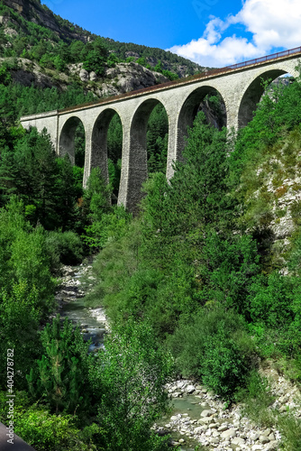 Old railway bridge, made of stone and arches, over the river Coulomp, with forest and mountains around, blue sky, Provence-Alpes-Côte d'Azur region, Alpes de Haute Provence, France