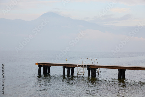 Pontile di legno vuoto su Lago di Garda con scaletta e montagne sullo sfondo photo