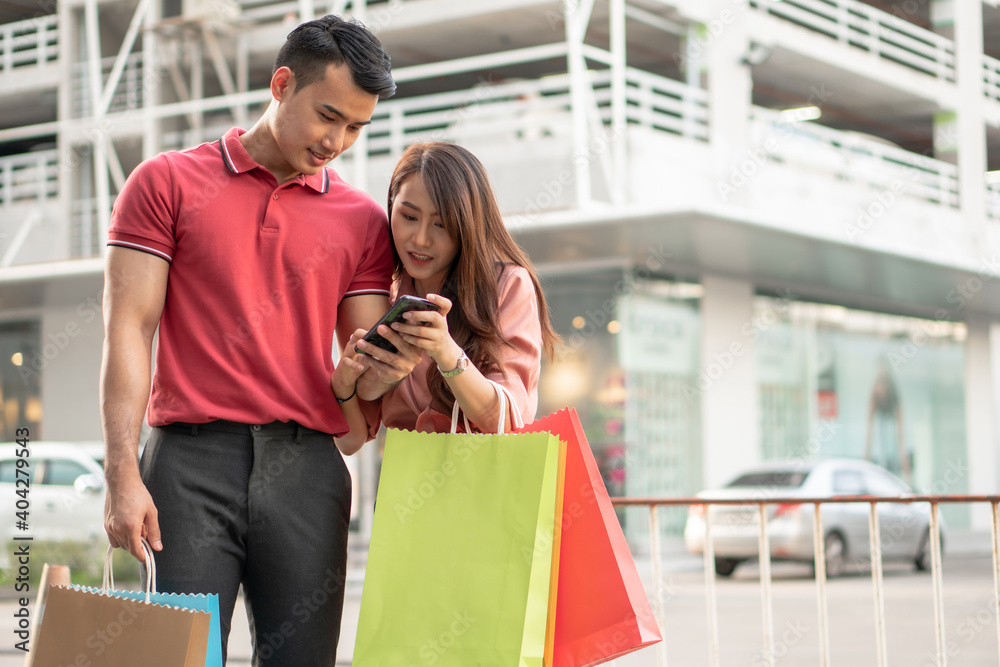 Happy young couple of shoppers walking in the shopping street towards and holding colorful shopping bags in hand and use a smartphone for check promotion. Concept of sale and Black Friday shopping Happy young couple of shoppers walking in the shopping street towards and holding colorful shopping bags in hand and use a smartphone for check promotion. Concept of sale and Black Friday shopping