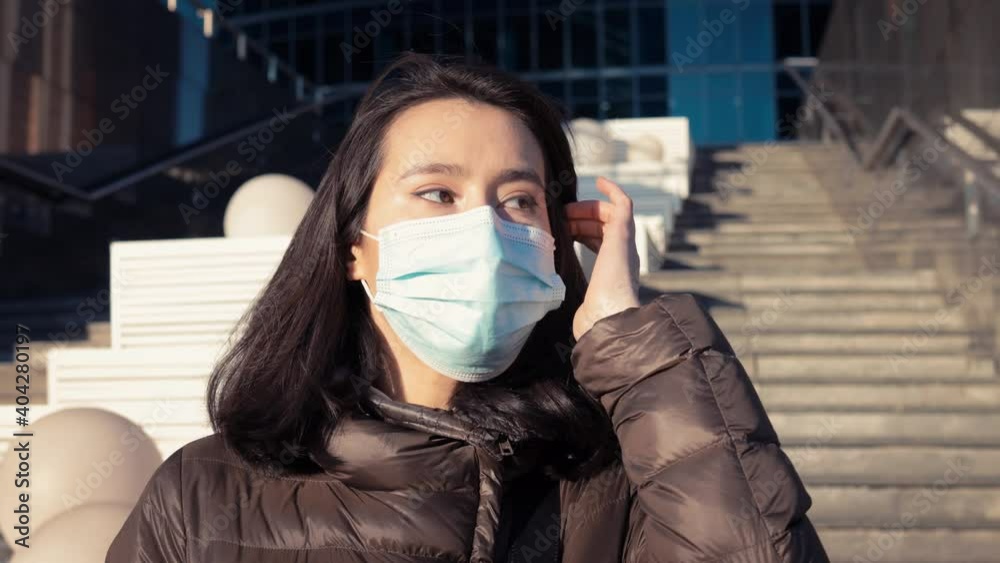 Young woman in protective mask, outside on background of business buildings steps. Pan shot right to left of a girl in medical protective mask portrait standing near big building Young woman in protective mask, outside on background of business buildings steps. Pan shot right to left of a girl in medical protective mask portrait standing near big building