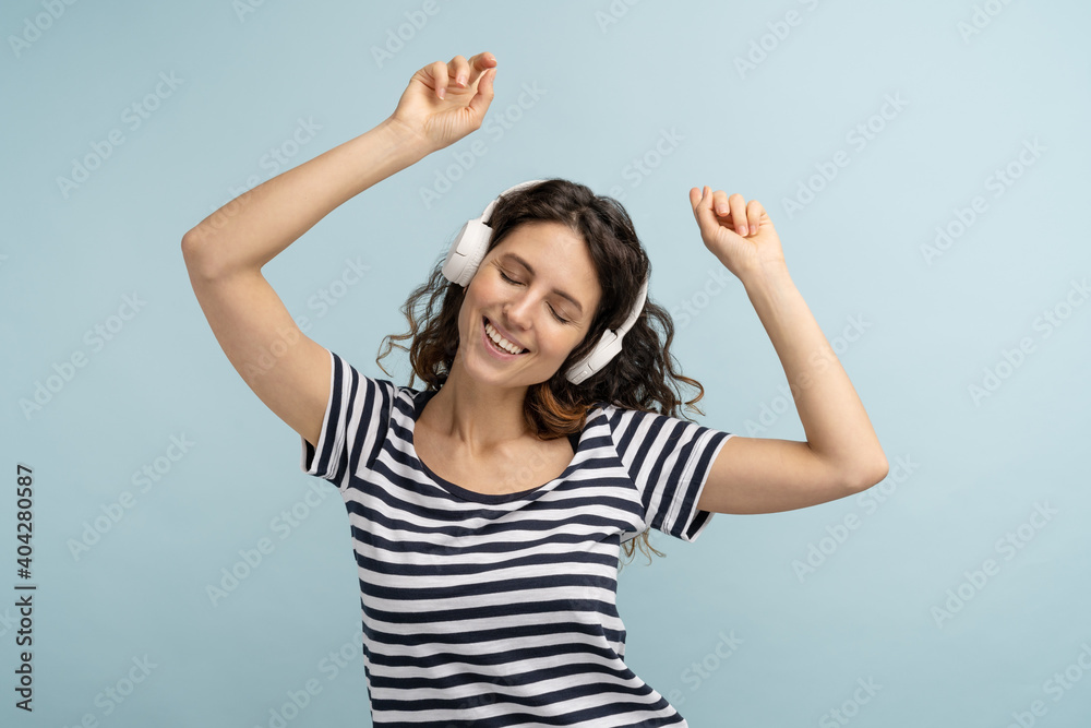 Energized happy caucasian woman in stripped t shirt sing along, wears wireless headphones, listens to music, dancing with hands up, has good mood, isolated on studio blue background Energized happy caucasian woman in stripped t shirt sing along, wears wireless headphones, listens to music, dancing with hands up, has good mood, isolated on studio blue background