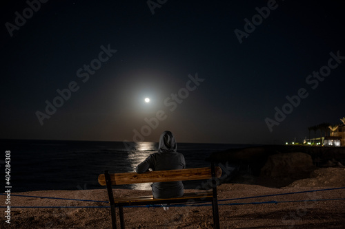 woman sits on a bench and looks at the rising moon over the sea at night