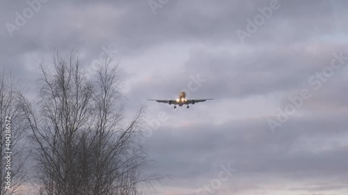 Airplane with flashing signal lights flies to land at Riga Airport under sky with grey clouds and bare tree on foreground