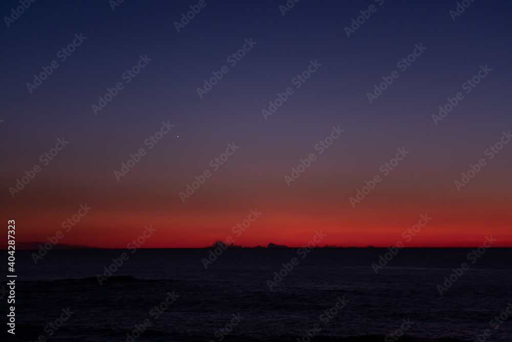 Vivid red blue gradient sky over the ocean at dusk with dark clouds on the horizon and star visible Vivid red blue gradient sky over the ocean at dusk with dark clouds on the horizon and star visible