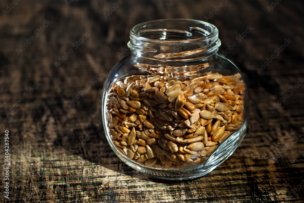 Sunflower seeds in a glass transparent jar. Nuts on an old shabby board. Jar on a brown wooden table close up. Dramatic contrasting light as an artistic effect. Sunflower seeds in a glass transparent jar. Nuts on an old shabby board. Jar on a brown wooden table close up. Dramatic contrasting light as an artistic effect.