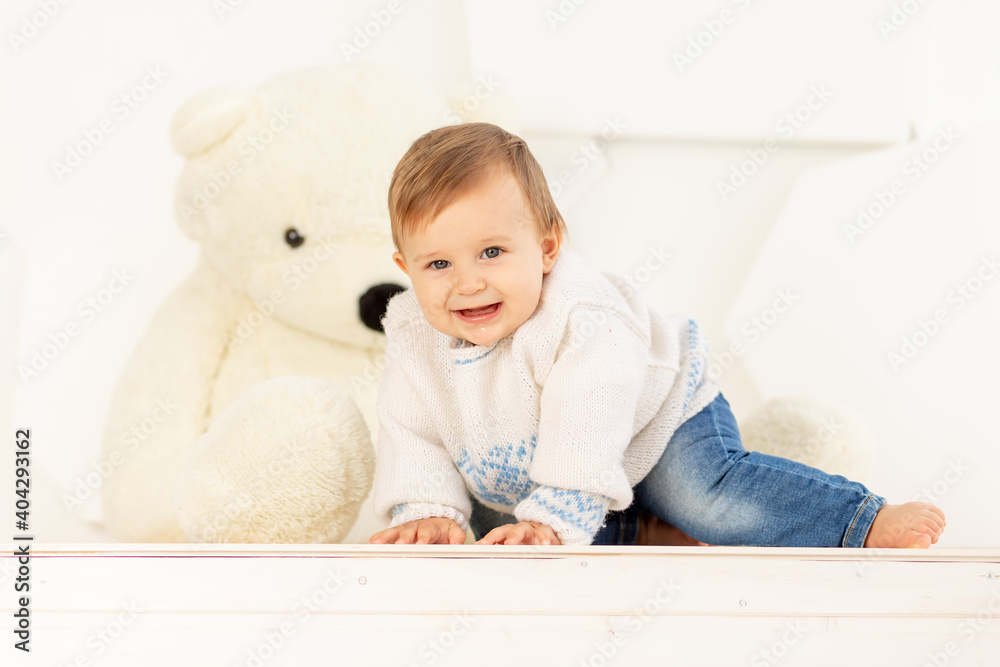 a happy little child six months old in a knitted warm jacket and blue jeans crawls at home near a large Teddy bear a happy little child six months old in a knitted warm jacket and blue jeans crawls at home near a large Teddy bear
