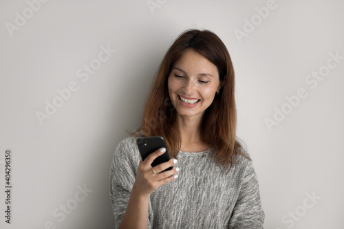 Smiling young red-haired Caucasian woman isolated on grey studio background use modern smartphone. Happy millennial female client or customer text message on cellphone. Technology concept.