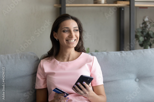 Smiling millennial indian Arabic woman sit on sofa at home shopping online on cellphone with credit card. Happy young ethnic female client buyer pay on internet use secure bank system on smartphone.