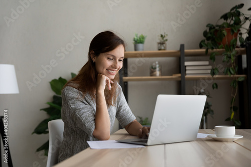 Smiling millennial Caucasian woman sit at desk at home office work distant on laptop. Happy young female look at computer screen browse surf wireless internet on gadget. Technology concept.