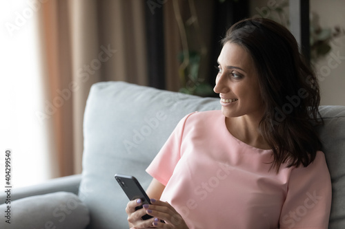 Happy millennial indian Arabic woman use smartphone look in distance window dreaming or thinking. Smiling young ethnic female relax at home browse cellphone texting, visualize or imagine.