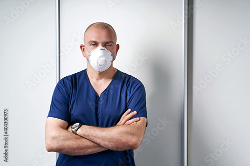 Photography Experienced male massage therapist masseur in his medical office standing by a massage table wearing professional uniform and disposable standard N95 filtering facepiece respirator face mask