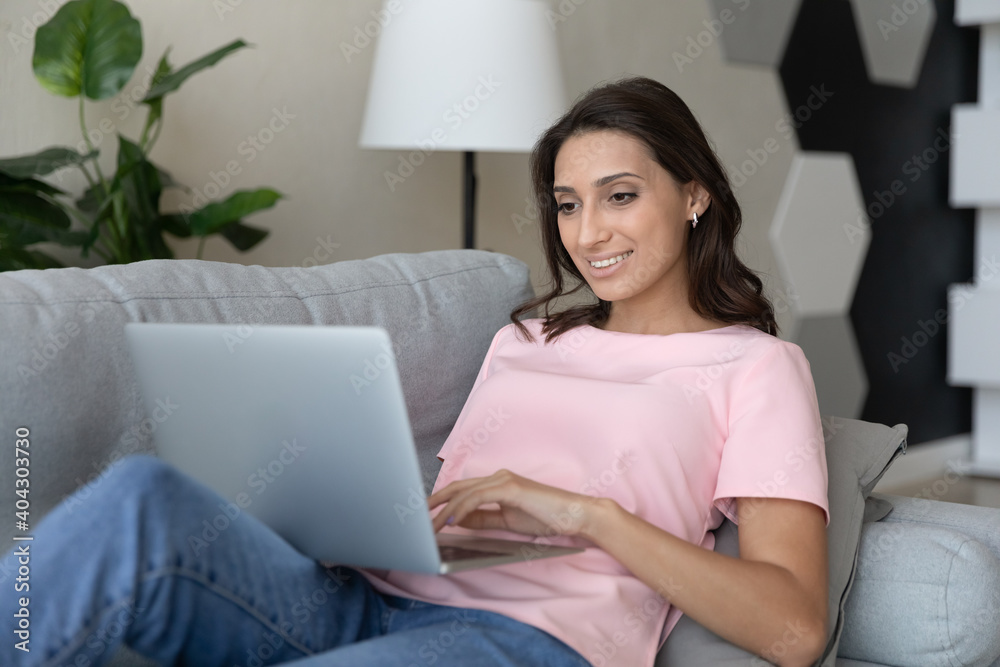 Smiling millennial indian Arabic woman relax on couch at home look at laptop screen work online. Happy young middle eastern female rest on sofa in living room browse surf on modern computer. Smiling millennial indian Arabic woman relax on couch at home look at laptop screen work online. Happy young middle eastern female rest on sofa in living room browse surf on modern computer.