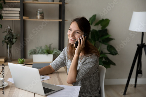Smiling young Caucasian woman sit at desk at home office use laptop talk on smartphone with client. Happy female employee or worker consult customer on cellphone call, work distant on computer.