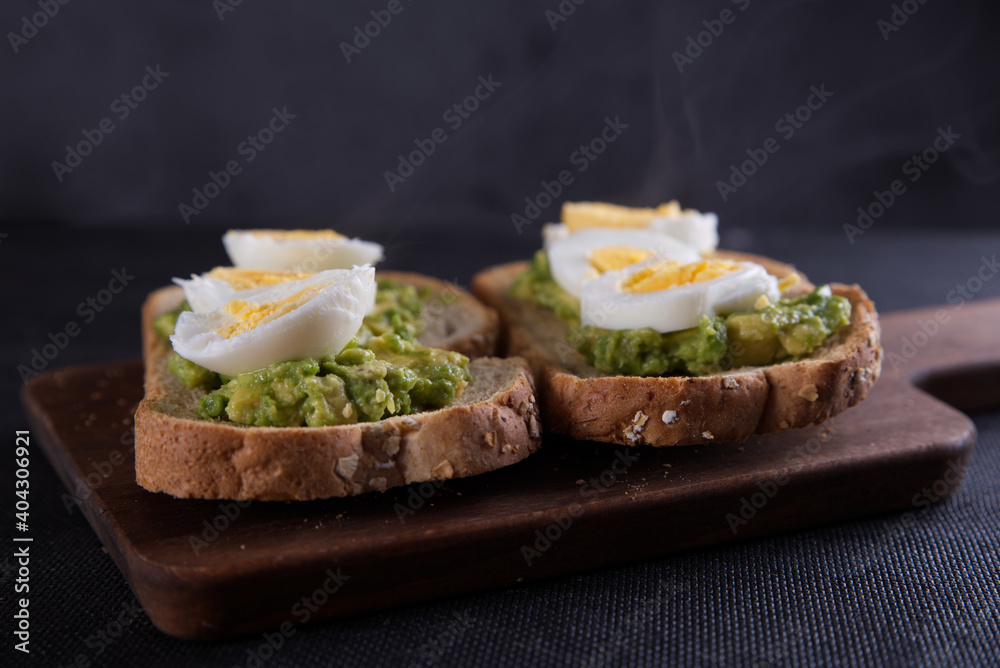 Sandwich prepared with oat toast, avocado and boiled egg, with dark backgrounds.