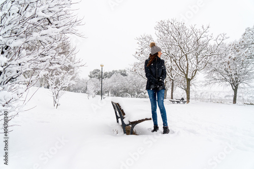 Chica joven en un parque nevado en un día de invierno con gorro de lana gris, jeans y cazadora negra