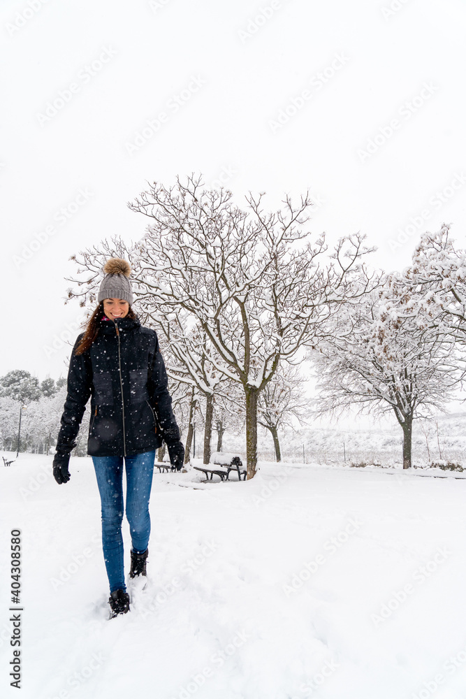 Chica joven en un parque nevado en un día de invierno con gorro de lana gris, jeans y cazadora negra