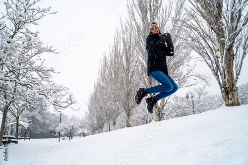 Chica joven en un parque nevado en un día de invierno con gorro de lana gris, jeans y cazadora negra photo