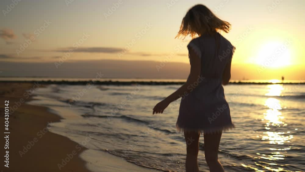 Cute young smiling woman running at sea on the beach around camera wearing blue summer dress during sunset Cute young smiling woman running at sea on the beach around camera wearing blue summer dress during sunset