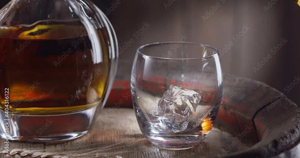 Jet of whiskey is poured into a glass with ice cubes, standing on a vintage whiskey barrel. A decanter with a drink stands nearby, a dark brown background.