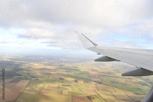 High view from airplane window of landscape of colorful farmland with green, light green and brown soil under white airplane wing, with blue sky white clouds background