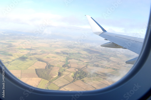 High view from airplane window of landscape of colorful farmland with green, light green and brown soil under white airplane wing, with blue sky white clouds background