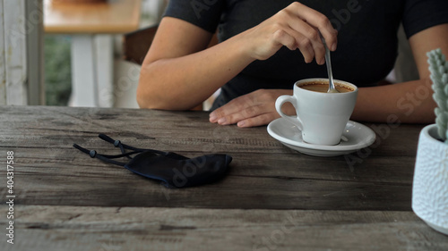uropean girl sits at a table in a public place with a cup of coffee and nearby to her on a wooden table is a black protective mask