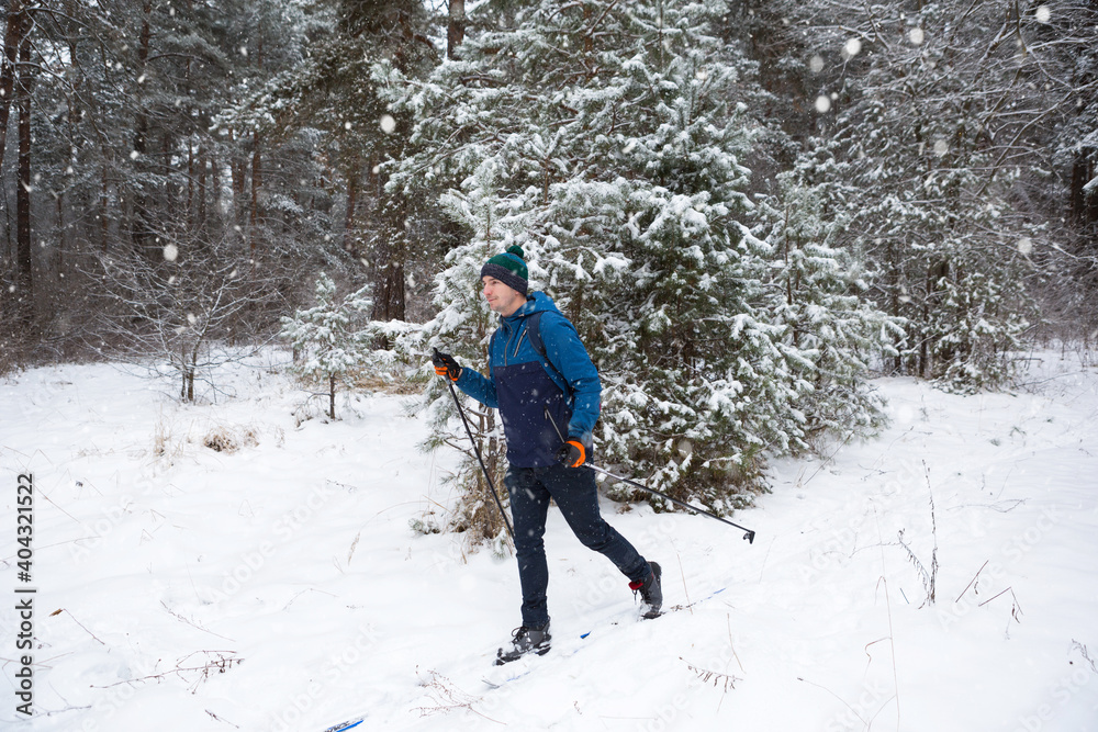Skier with a backpack and hat with pompom with ski poles in his hands on background of a snowy forest. Cross-country skiing in winter forest, outdoor sports, healthy lifestyle, winter sports tourism. Skier with a backpack and hat with pompom with ski poles in his hands on background of a snowy forest. Cross-country skiing in winter forest, outdoor sports, healthy lifestyle, winter sports tourism.