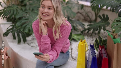Young woman have a rest taping of smartphone in shopping mall. Attractive blonde female sitting in the mall with colorful bags. indoor shopping. Happy girl texting, look at the side and smiling.