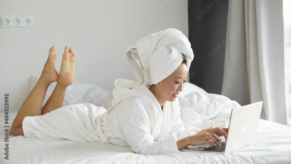 beautiful young Chinese woman working with a laptop, lying in bed at home, working remotely online due to the coronavirus pandemic. beautiful young Chinese woman working with a laptop, lying in bed at home, working remotely online due to the coronavirus pandemic.