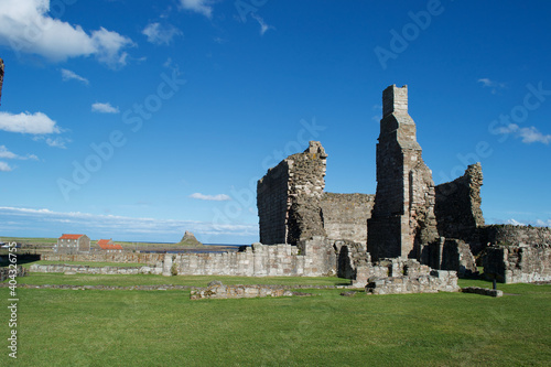 Ruins of a large stone building by the coast on a sunny day. Tall chimney stack standing amongst crumbling walls, beneath blue skies. (Lindisfarne, Holy Island, Northumberland in background)