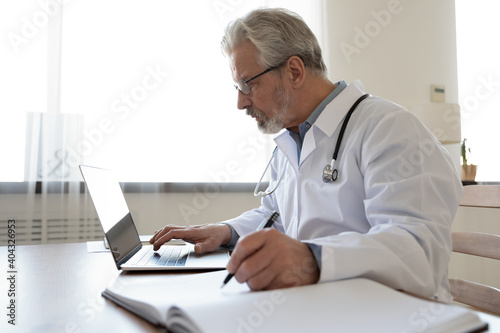 Side view concentrated 60s mature senior male doctor in glasses working on computer, writing notes in paper registry album, managing patient's appointments or disease treatments at hospital office.