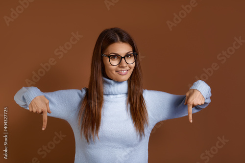 Look there. Charming carefree caucasian woman wearing glasses and light blue sweater points down with both index fingers, has broad interested smile, isolated over brown background.
