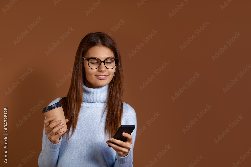 Cheerful smiling caucasian woman wearing glasses and light blue sweater holds coffee in one hand and mobile phone in the other, reading good news, chatting with friends, isolated on brown background. Cheerful smiling caucasian woman wearing glasses and light blue sweater holds coffee in one hand and mobile phone in the other, reading good news, chatting with friends, isolated on brown background.
