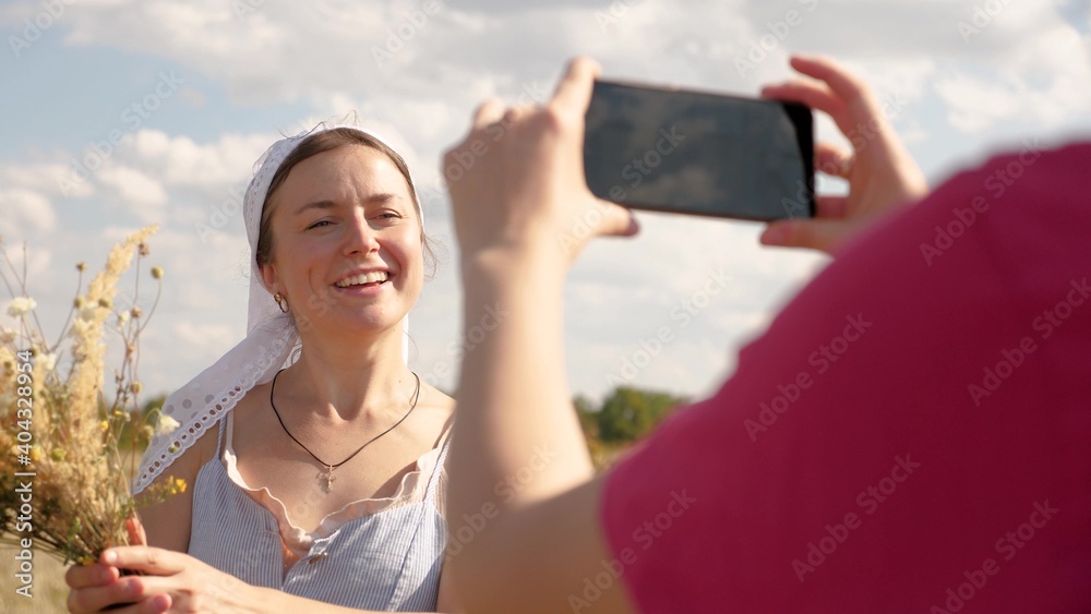 Two happy girlfriends spend time outdoors on beautiful sunny day. Online video of beautiful woman in meadow. Ecotourism. Girl blogger in field with flowers. Young woman is photographed on smartphone