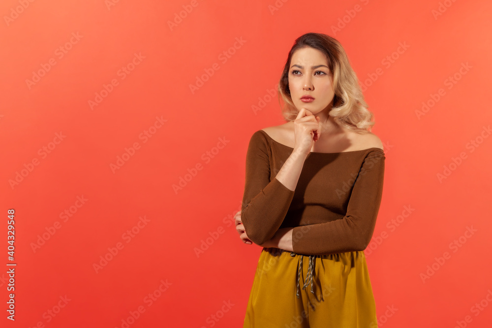 Portrait of a young woman in brown blouse pondering with a serious expression. She touching her chin and thinking with thoughful face, looking away. Indoor studio shot isolated on red background Portrait of a young woman in brown blouse pondering with a serious expression. She touching her chin and thinking with thoughful face, looking away. Indoor studio shot isolated on red background