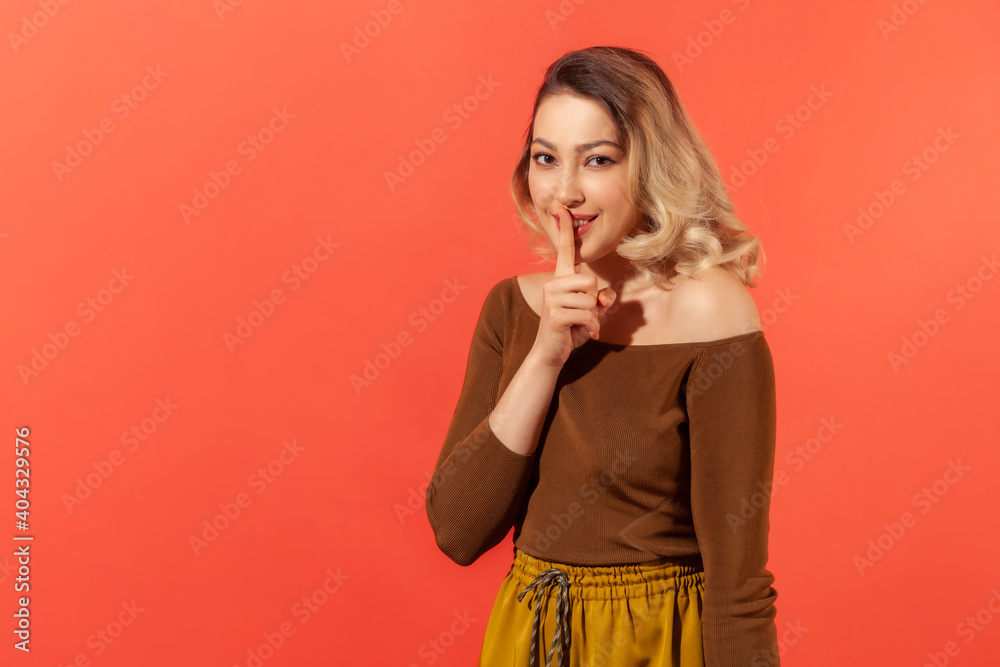 Shh, it's big secret. Beautiful cunning woman with blonde hair in brown casual blouse smiling, showing gesture secret sign with finger near her lips. Indoor studio shot isolated on red background Shh, it's big secret. Beautiful cunning woman with blonde hair in brown casual blouse smiling, showing gesture secret sign with finger near her lips. Indoor studio shot isolated on red background
