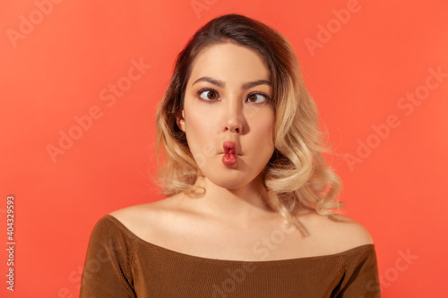 Portrait of funny crazy young beautiful blonde womanl in brown blouse standing and looking with crossed eyes and fish lips. Indoor studio shot isolated on red background